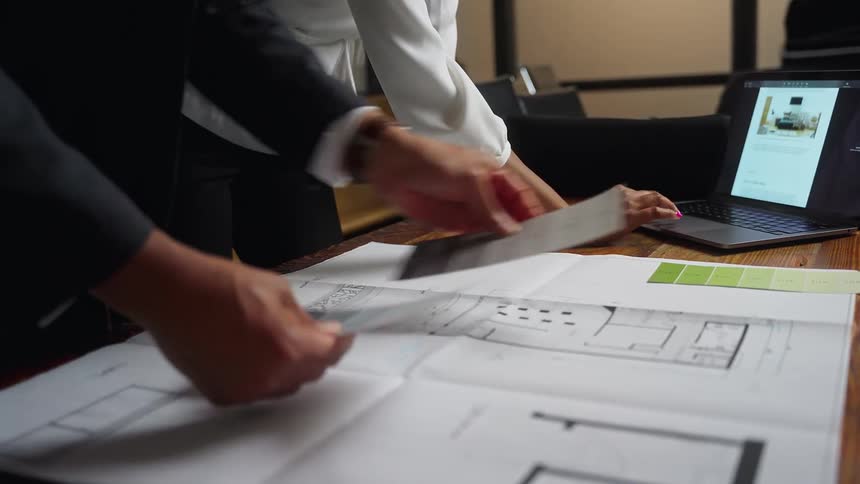 Two people are leaning over a desk with a laptop computer and floor plans for a room while looking at paint color swatches