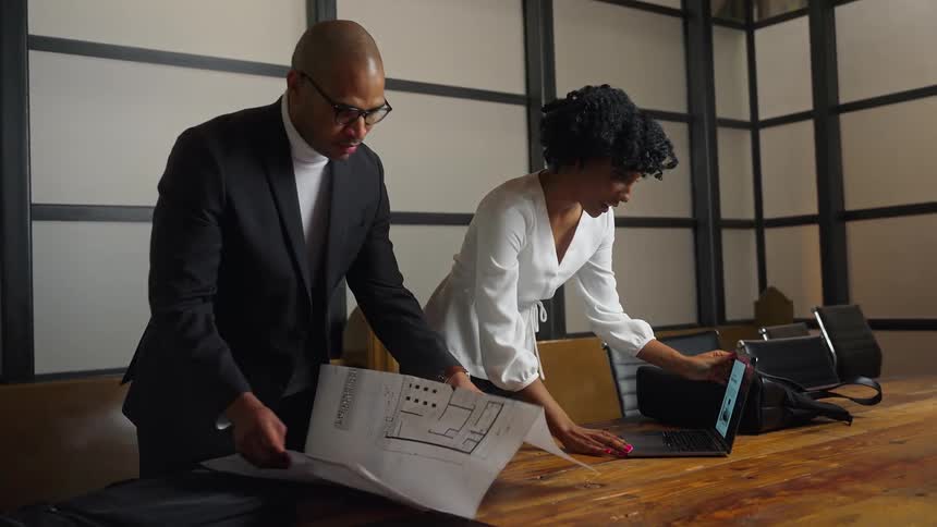 A woman unpacks her laptop while a coworker unfolds paper floor plans for their meeting
