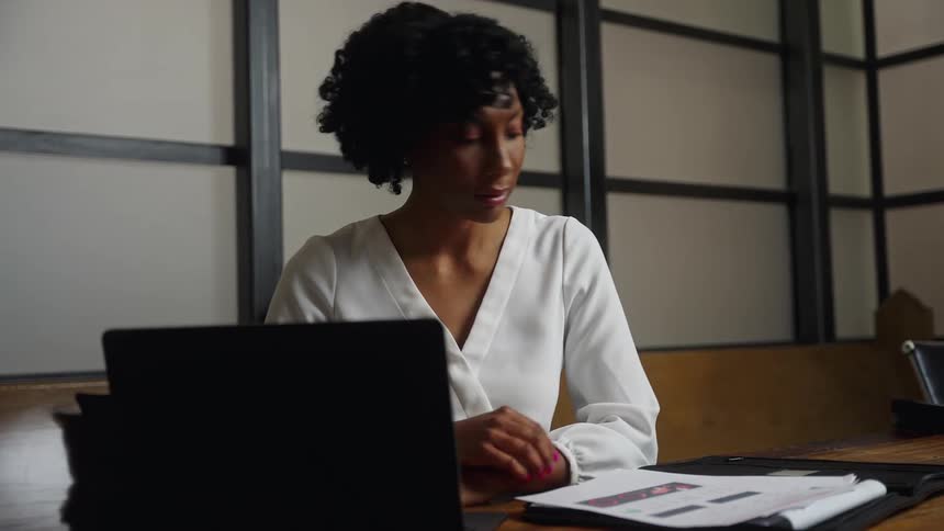 A woman sitting in an office conference room looking through pages of information and typing on a laptop
