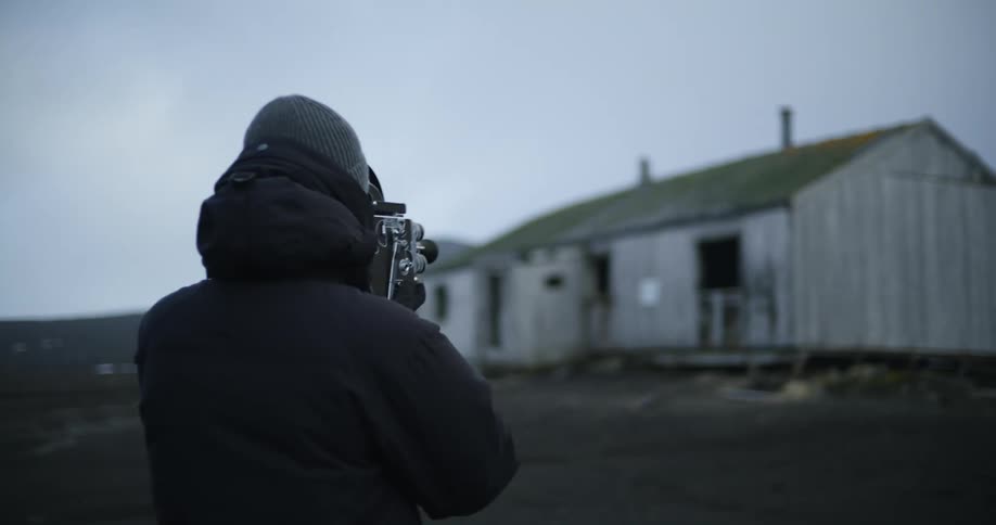 Camera operator wearing coat pans film camera over abandoned farmhouse sitting near mountain