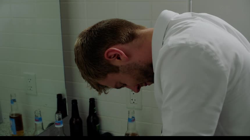 A man in a dress shirt lean forward over a bathroom sink that is littered with beer bottles