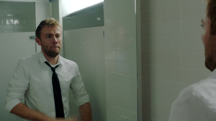 A man is in a public bathroom with beer bottles on the sink counter as he rubs his face and looks at himself in the mirror