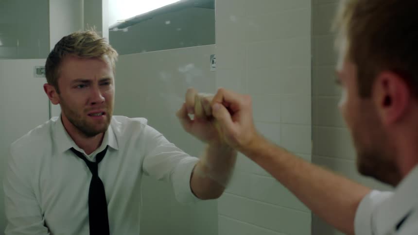 A man in a dress shirt stares at his reflection in a bathroom mirror as hes overcome with emotion