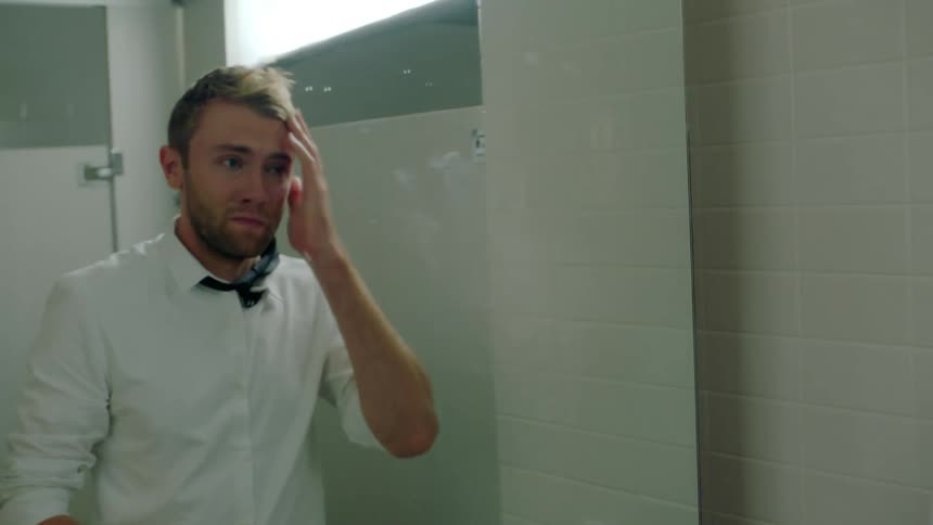 A man is in a public bathroom and he moves his necktie over his shoulder and wipes his face and hair with his hand as he looks in the mirror