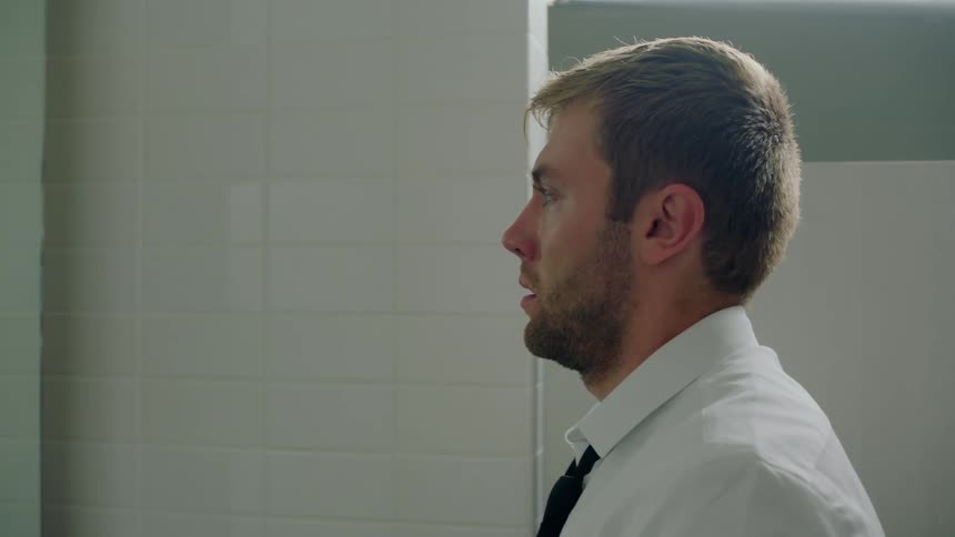 Man looks down sadly after fixing his tie while standing in tiled bathroom