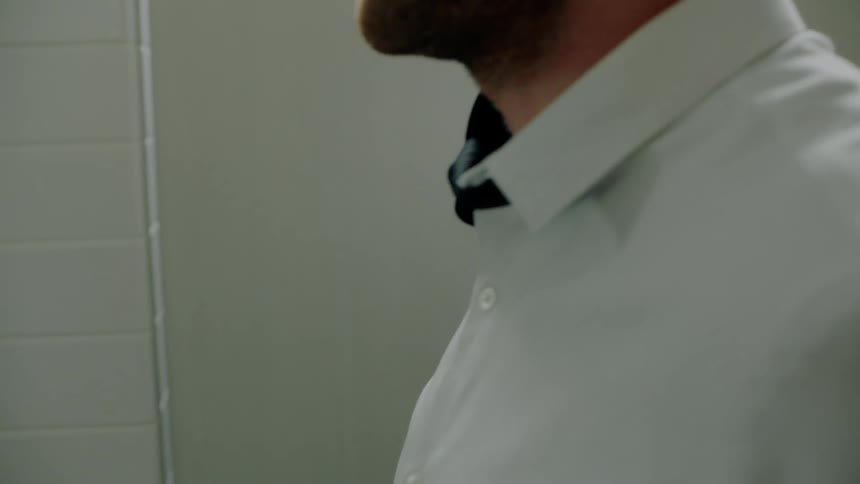 A man is in a small bathroom and he moves his necktie over his shoulder