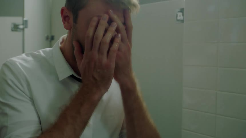 A man is looking into a bathroom mirror appearing distraught and splashes water onto his face and pats his hair with his hand