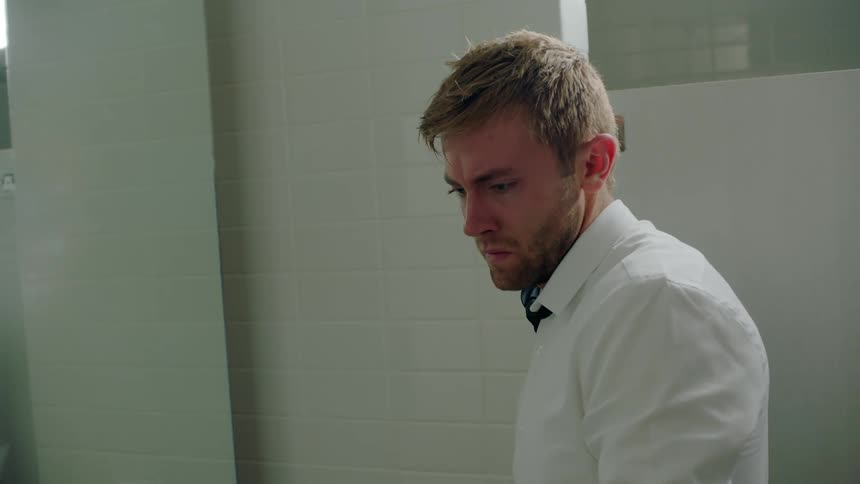 A man is in a bathroom and he sets his beer on the sink counter near empty beer bottles and pulls his necktie off his shoulder and walks away