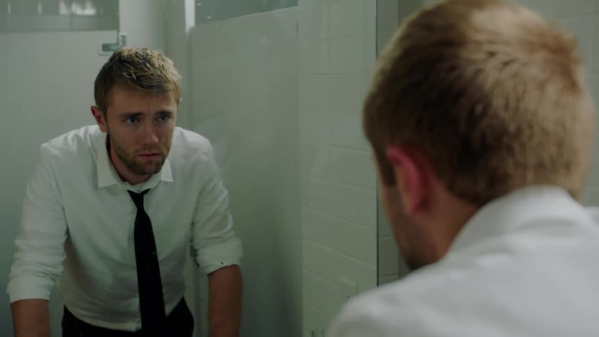 A man in an office bathroom looks up at his reflection shaking his head looking distraught