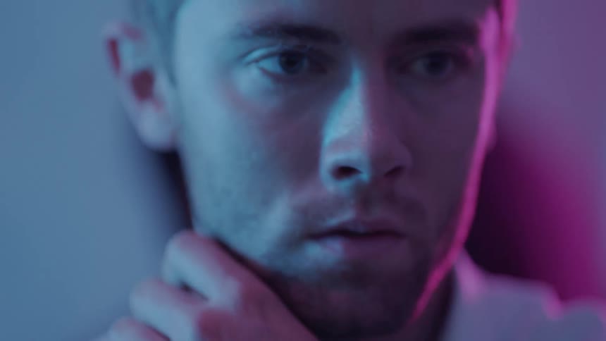 A man is in a room looking around while rubbing his neck and there are other people moving behind him