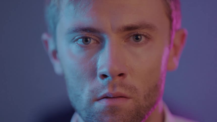 Man stands staring under colored lighting as people walk behind him at party