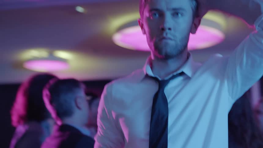 Man stands at party under colored lighting holding a beverage bottle and thinking