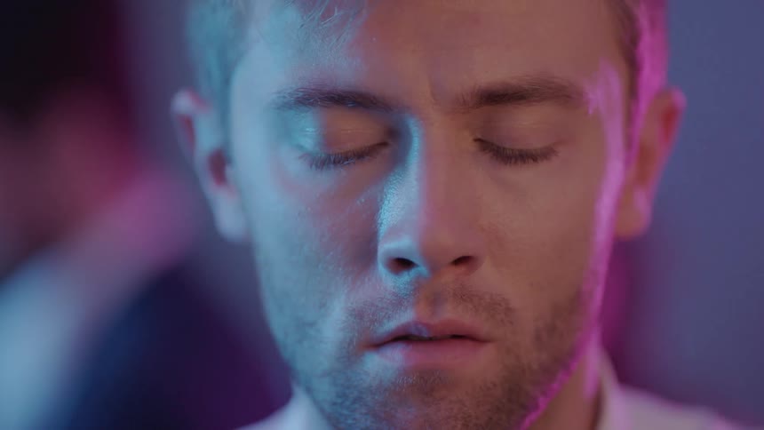 A man whose face is illuminated with light from a screen closes his eyes in despair