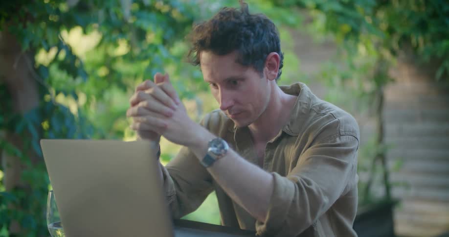 A man is sitting outside at a table typing on a laptop computer with his phone sitting on the table and he stops to rub his face with his hands