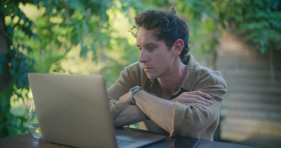 A man sitting at a table on the patio taps his fingers as he thinks then begins to type on a laptop