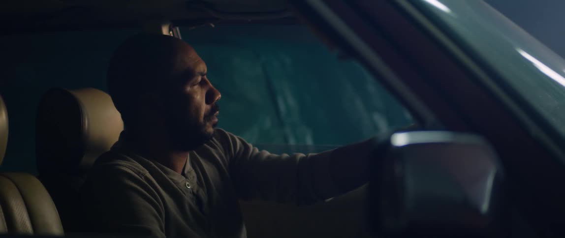 A man is sitting in the drivers seat of a parked automobile with the hood up and he looks tired as he holds the steering wheel