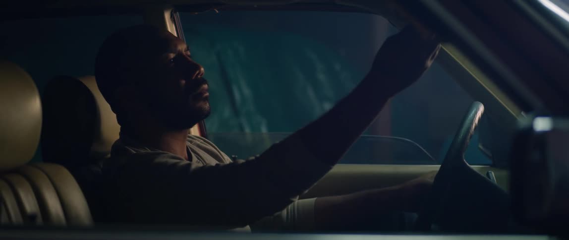 A man is sitting in the drivers seat of a parked automobile and he looks in the rear view mirror
