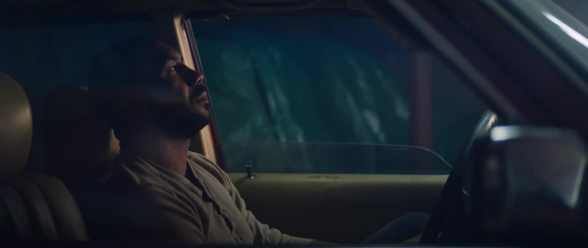 A man sits in his car at night and leans his head back on the head rest
