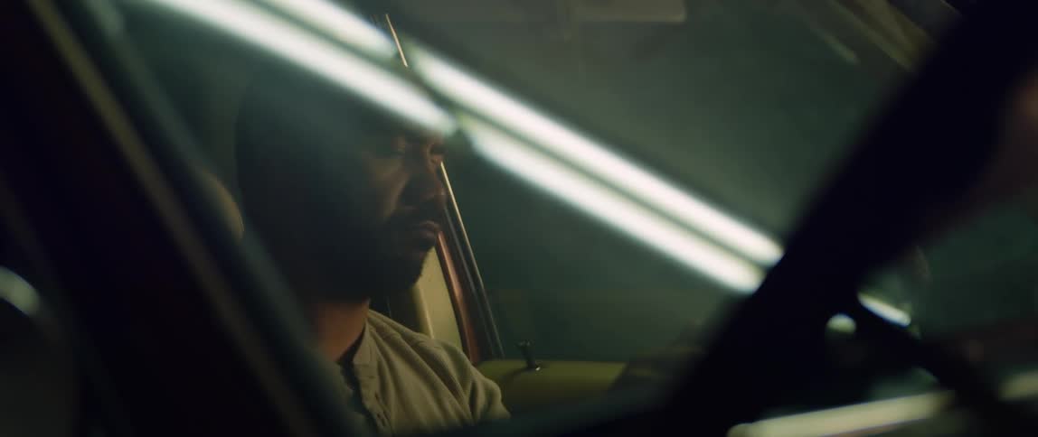 A man is sitting in the drivers seat of an automobile parked in a garage as a light shines across the window