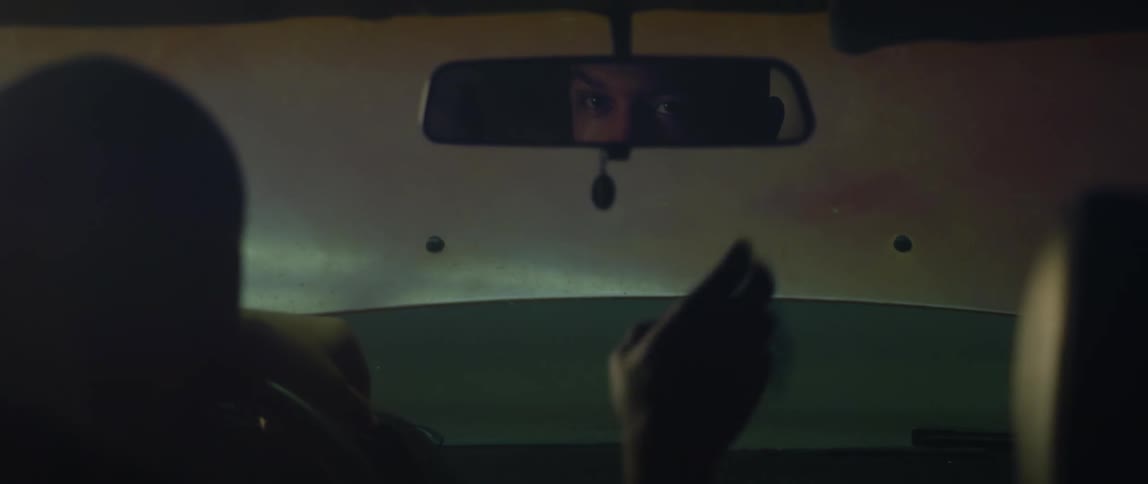 A man sitting in a parked car reaches up and adjusts the rearview mirror