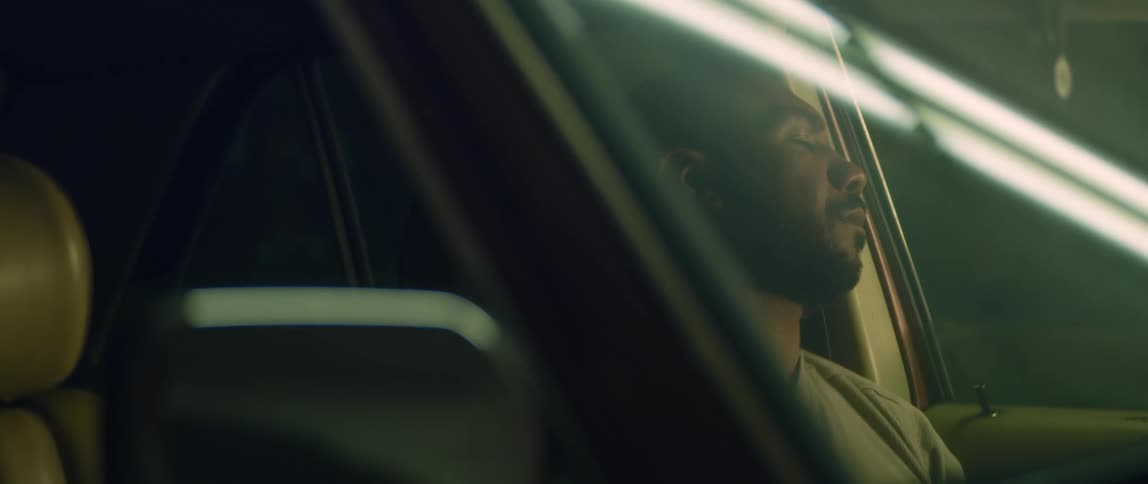A man is sitting in the drivers seat of an automobile parked in a garage as a light shines across the windshield