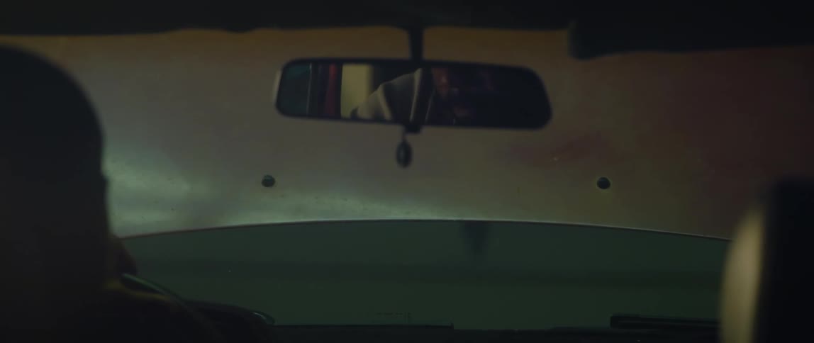 A man sits in his car with the hood up and adjusts the rearview mirror and looks in it