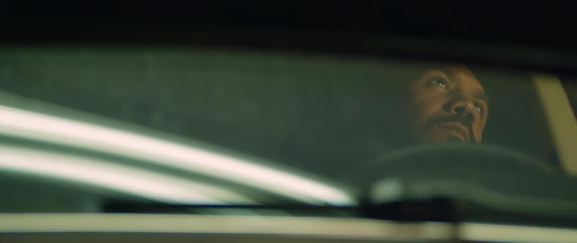 A man sits in his car at night with his eyes closed and his head leaning against the seat and then opens his eyes and looks around
