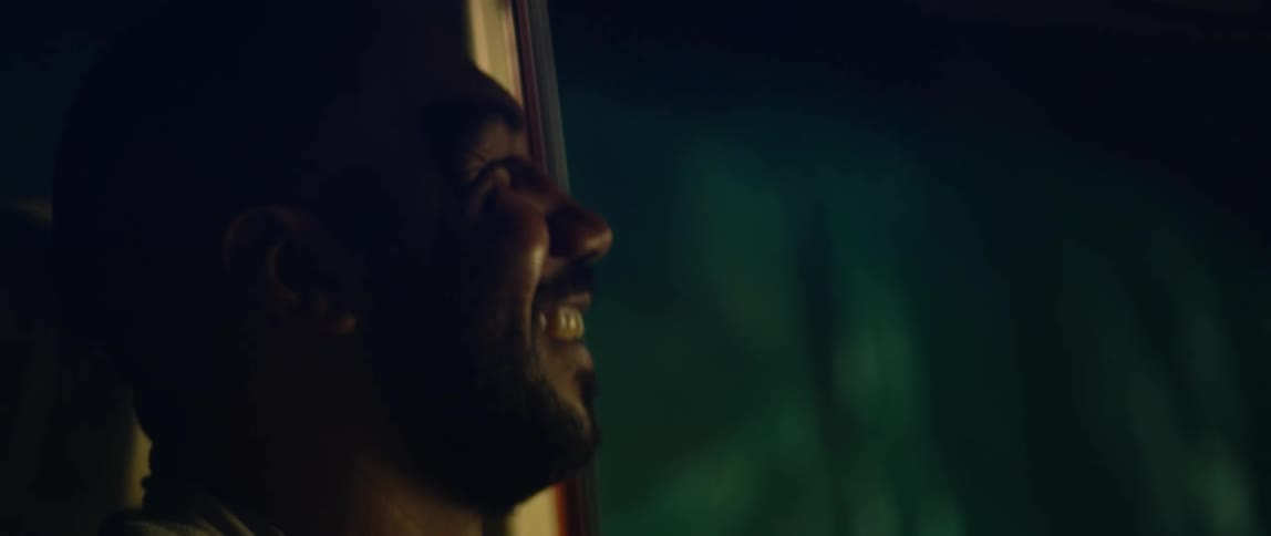A man sitting in his car with head tilted as if listening begins to smile and laugh