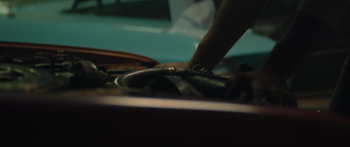 A man leaning on and looking at a car engine reaches to repair a part