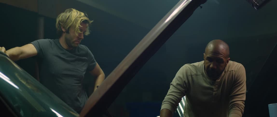 A man leans against a car with the hood up and another man comes and looks under the hood