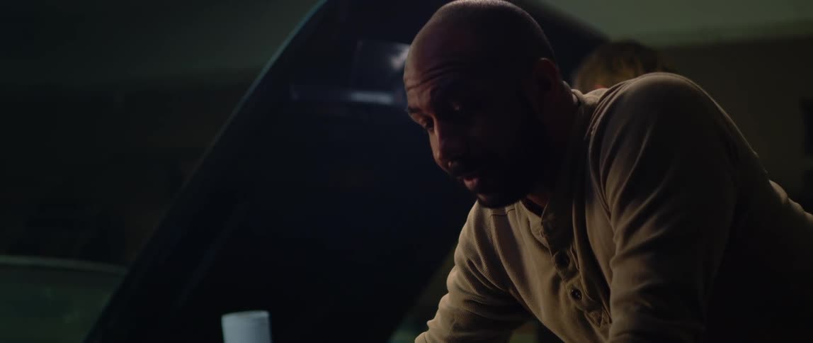 A man is working under the hood of a car and he speaks to another man working on a car behind him