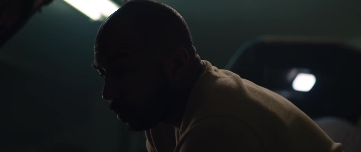 A man looks very tired as he works under the hood of an automobile in a dark garage with a bright ceiling light