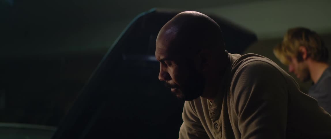 A man waits next to a car with a hood open as a mechanic works under the hood