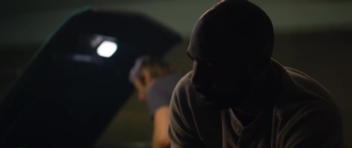 A man is looking under the hood of a car shaking his head and turns to the man working on a car behind him
