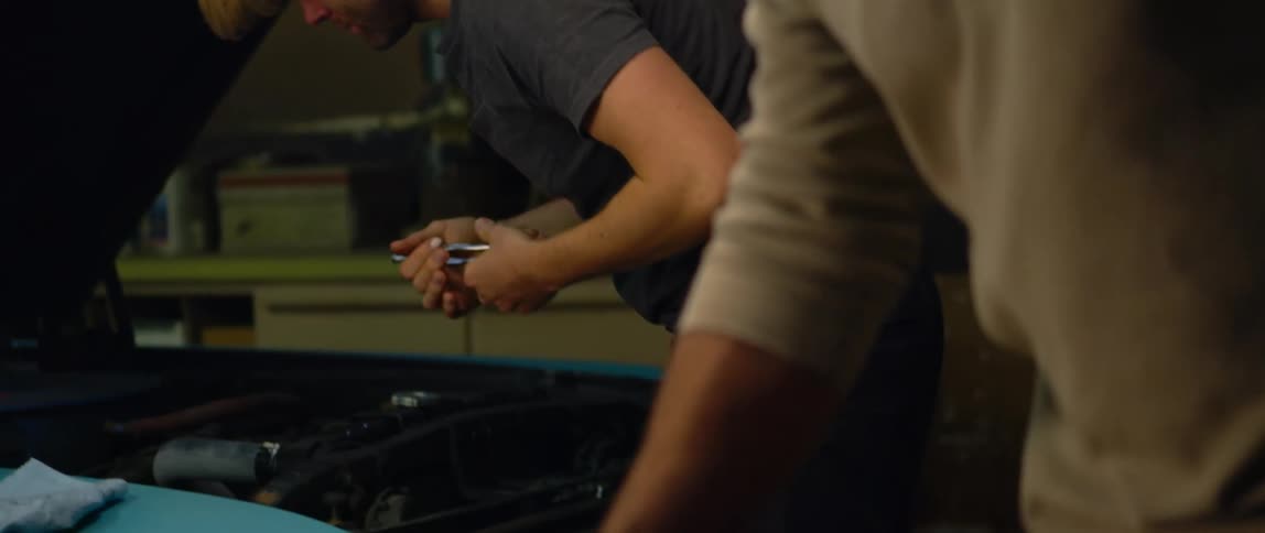 A man hands a wrench to a mechanic as he works under the hood of a car
