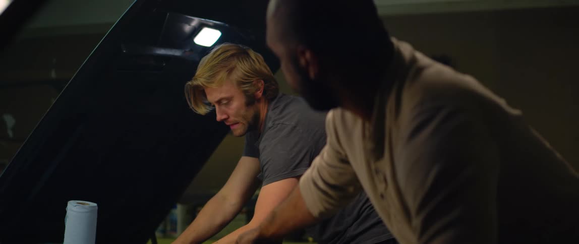 A man is working under the hood of a car and the man working on a car next to him hands him a wrench