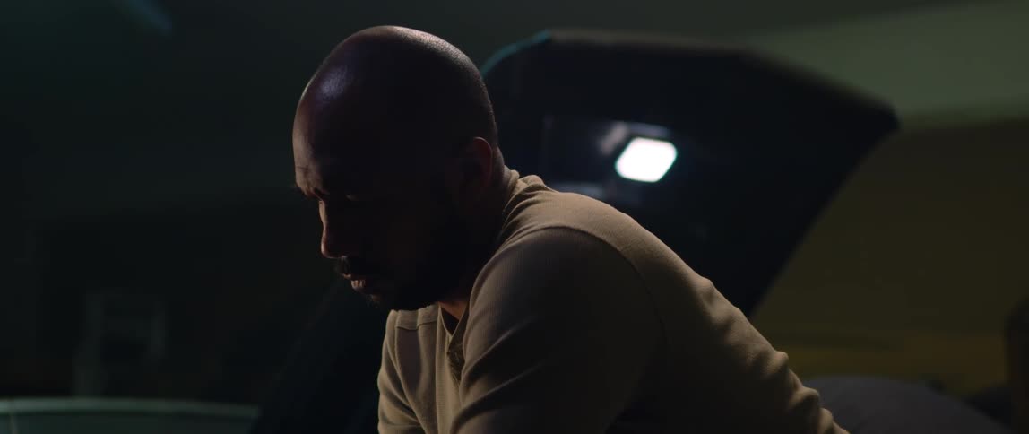 A man looking down at a car engine while a mechanic works under the hood of a car behind him