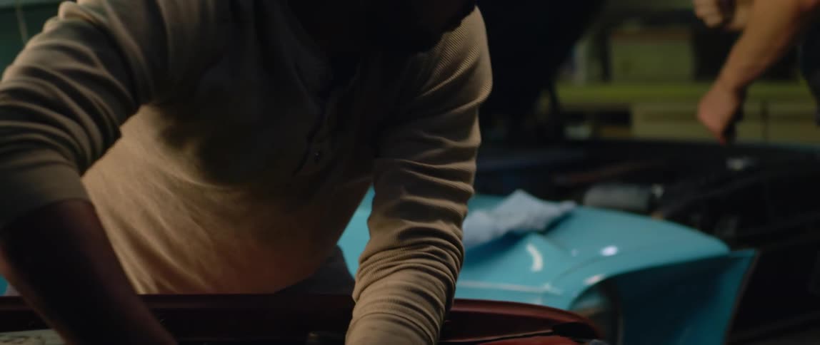 A mechanic is working under the hood of a car and he hands pliers to a man working on a car next to him