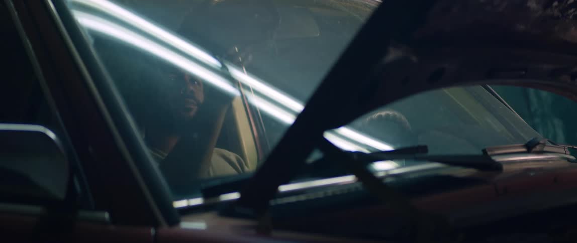 A man sits in his parked car with the hood up in a garage