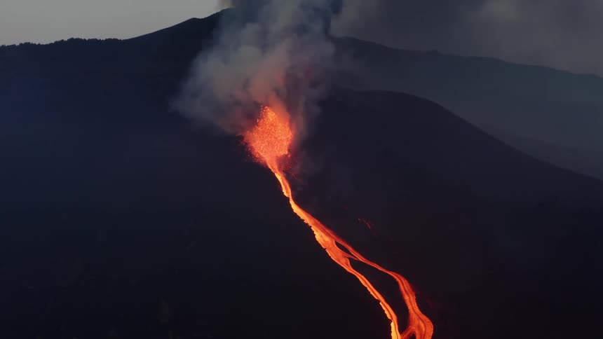 Bright orange lava flows in streams down a slope while a volcano erupts and smoke billows