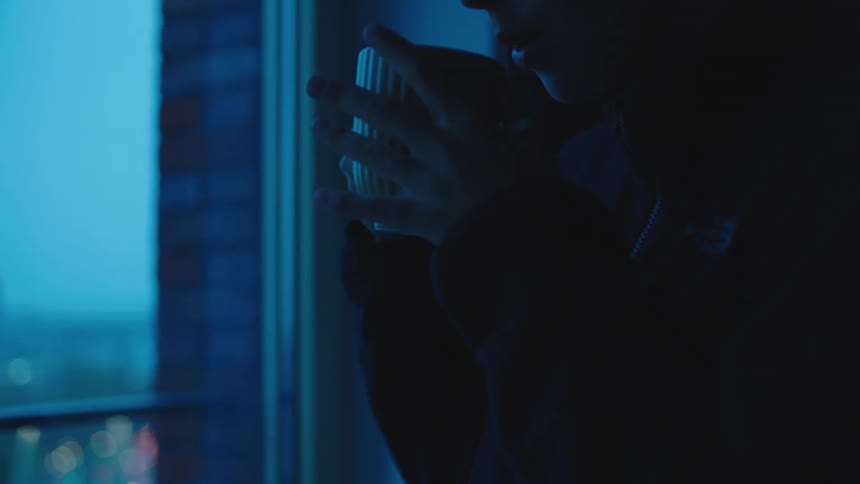 A woman walks toward her balcony doors and sips her hot mug while looking out at the city