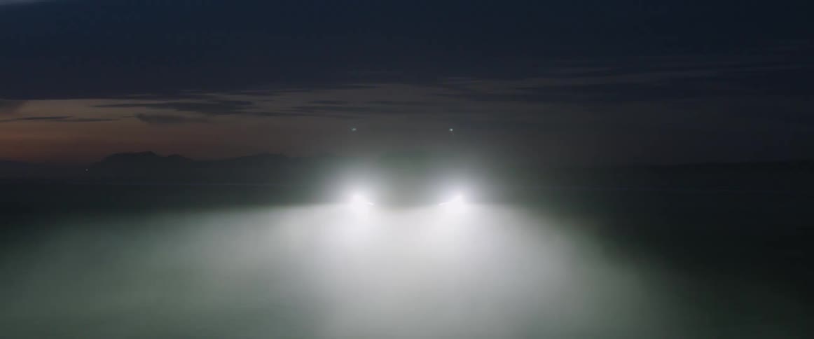 The headlights on an automobile illuminate the fog as it travels on a snow covered road and there are mountains in the distance