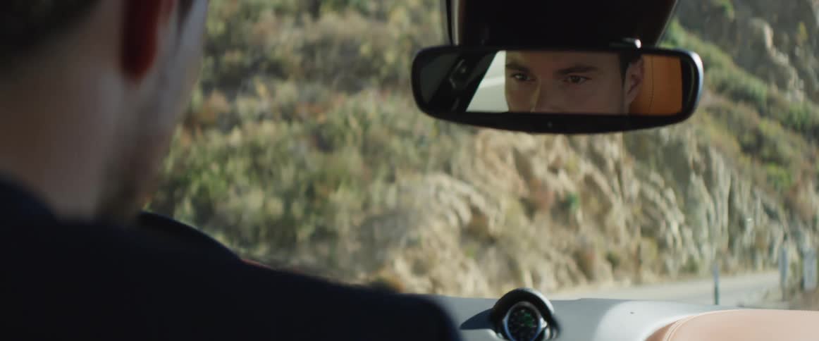 A mans face is seen in the rearview mirror as he drives an automobile traveling on a road alongside a mountain