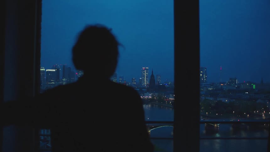 A woman in a dark room opens a curtain to reveal the skyline of a city at night