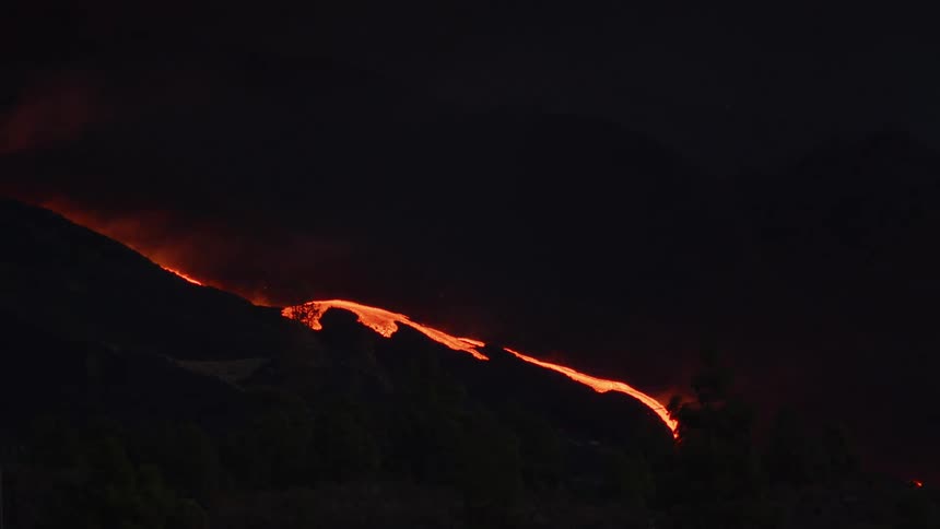 Bright hot lava flows down a slope with smoke and steam billowing at night