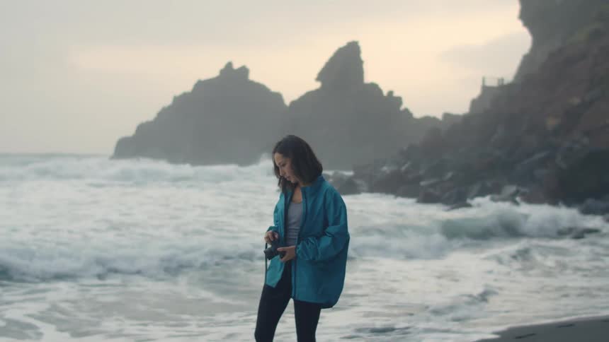 A female photographer walks along a beach while carrying a camera on a cloudy evening