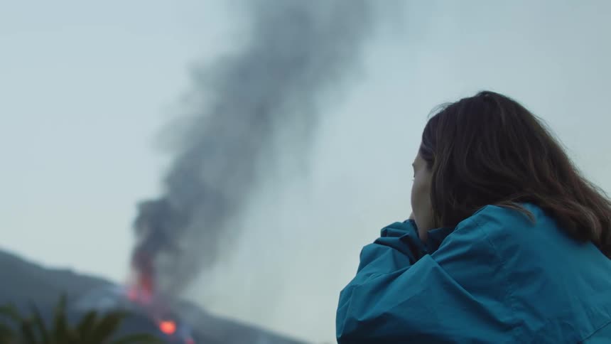 Smoke and flames rise from a fire on a mountain as a woman lifts her camera and takes a picture