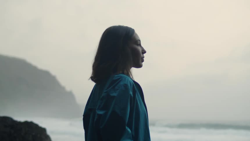 Woman with wavy hair and blue raincoat looks around on the beach next to the wavy ocean