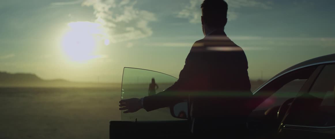 A woman is standing in the desert next to an automobile looking at the sun shining brightly over the mountains and a man gets out of a car parked nearby