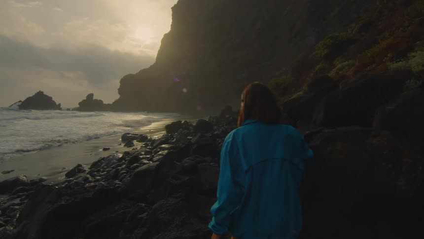 A woman is walking near rocks and she walks toward a beach as the waves roll in and she looks up at the sky